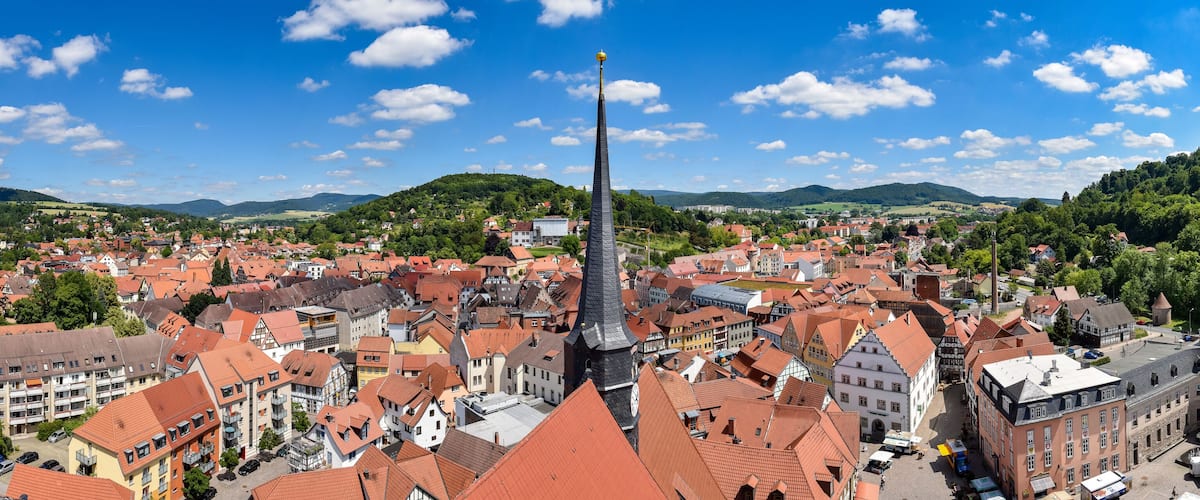 A town with red-roofed buildings, a prominent church tower, and green hills in the background.