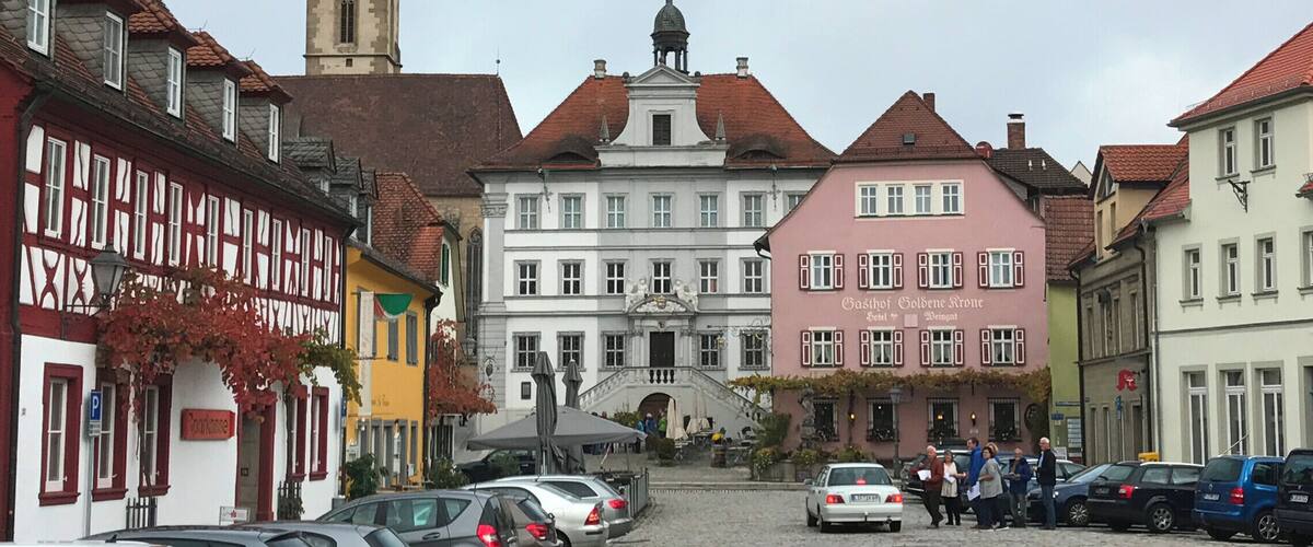 A cobblestone street lined with historic buildings, a church tower, and parked cars.