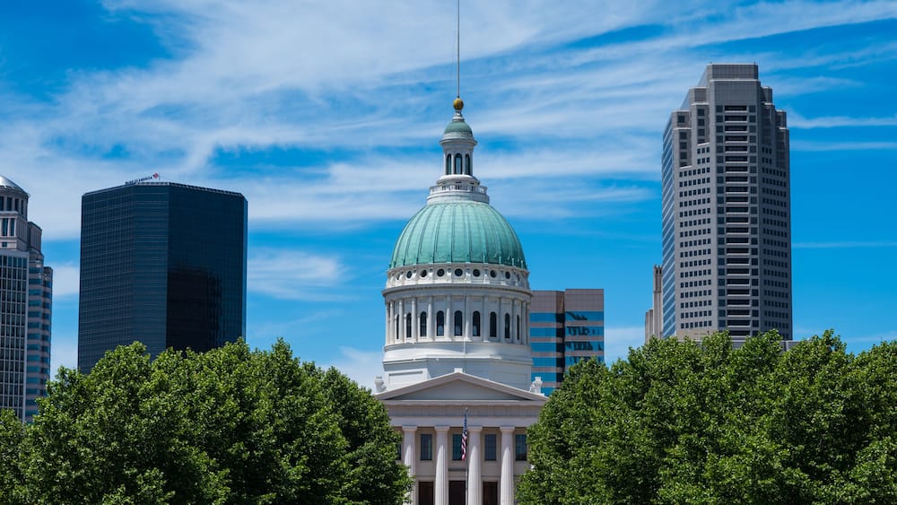 A cityscape with a prominent building featuring a green dome and classical architecture, surrounded by modern skyscrapers.