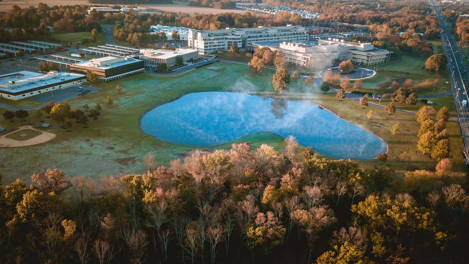 An aerial view of a campus with multiple buildings, a large pond, and surrounding wooded areas.
