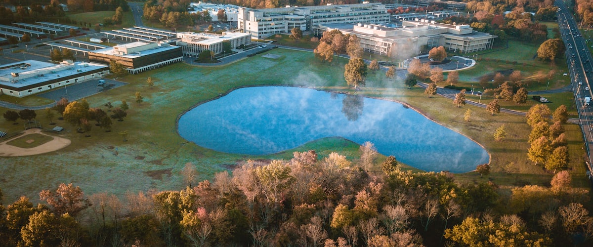 An aerial view of a campus with multiple buildings, a large pond, and surrounding wooded areas.