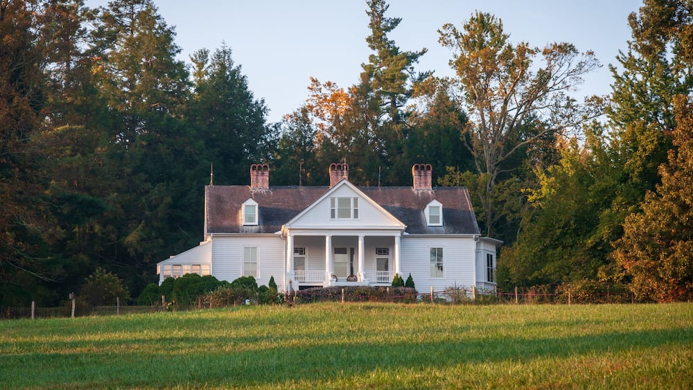 A white house with a porch, surrounded by a grassy field and trees.