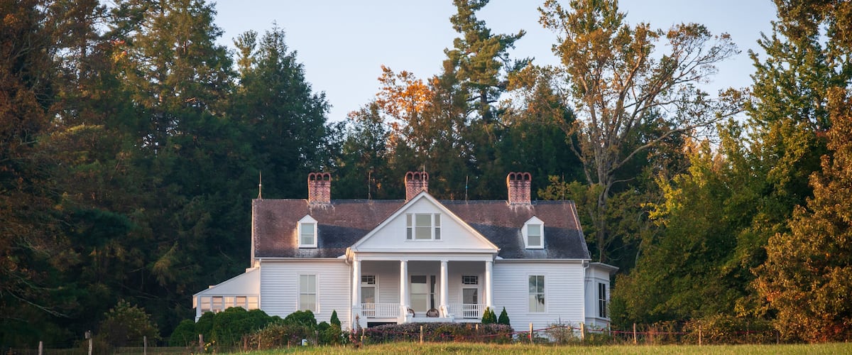 A white house with a porch, surrounded by a grassy field and trees.