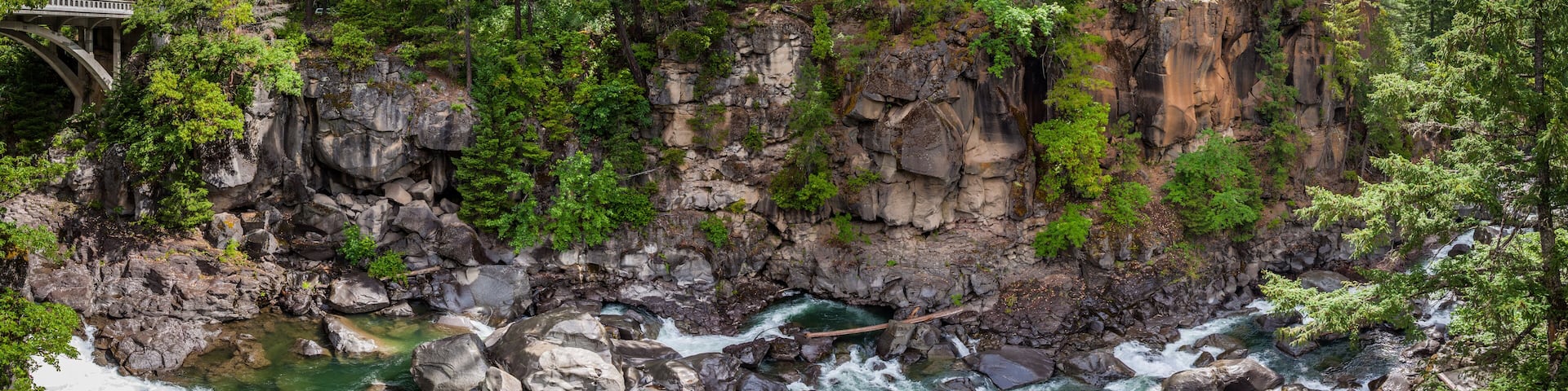 Panorama of a bridge on the left with a creek running between boulders in a narrow steep canyon in a dense forest near the Rogue River in Oregon