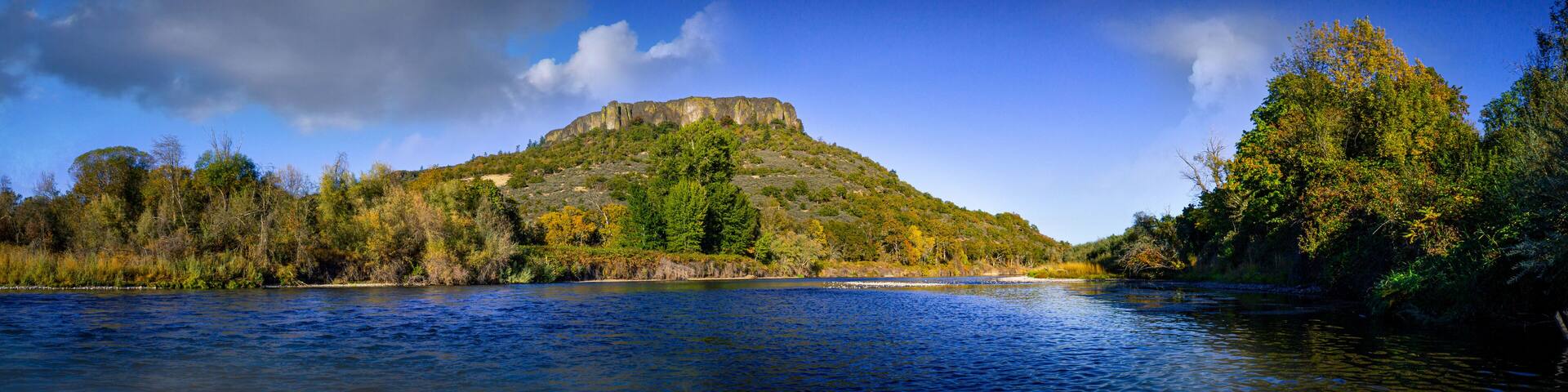 A river with a forested hill and a rocky peak in the background.