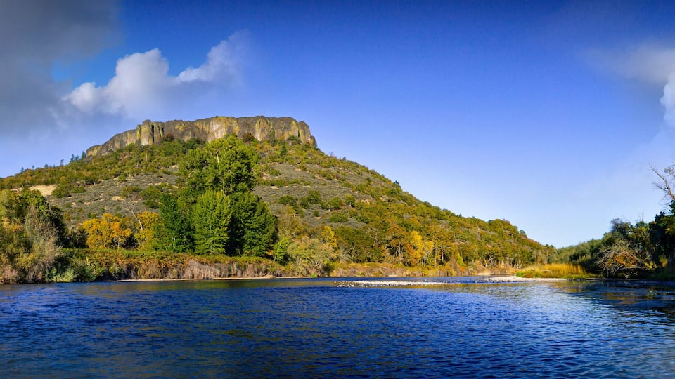 A river with a forested hill and a rocky peak in the background.