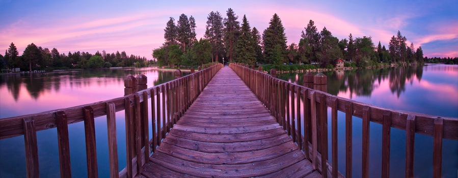 Un muelle de madera se adentra en un lago tranquilo, rodeado de árboles frondosos y un cielo colorido.
