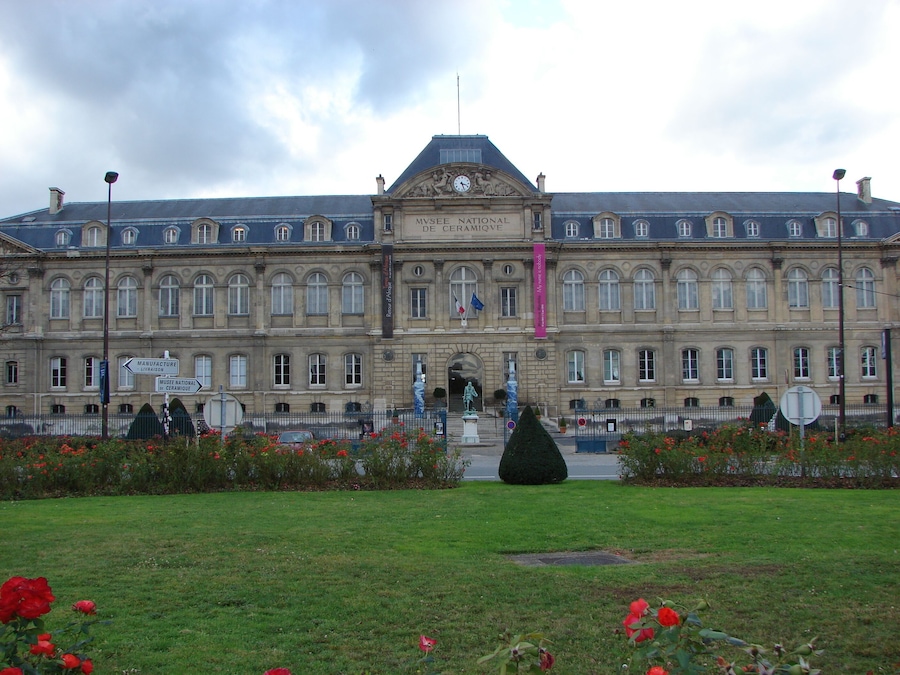 A large historic building with a clock tower, surrounded by a well-maintained garden with red flowers.