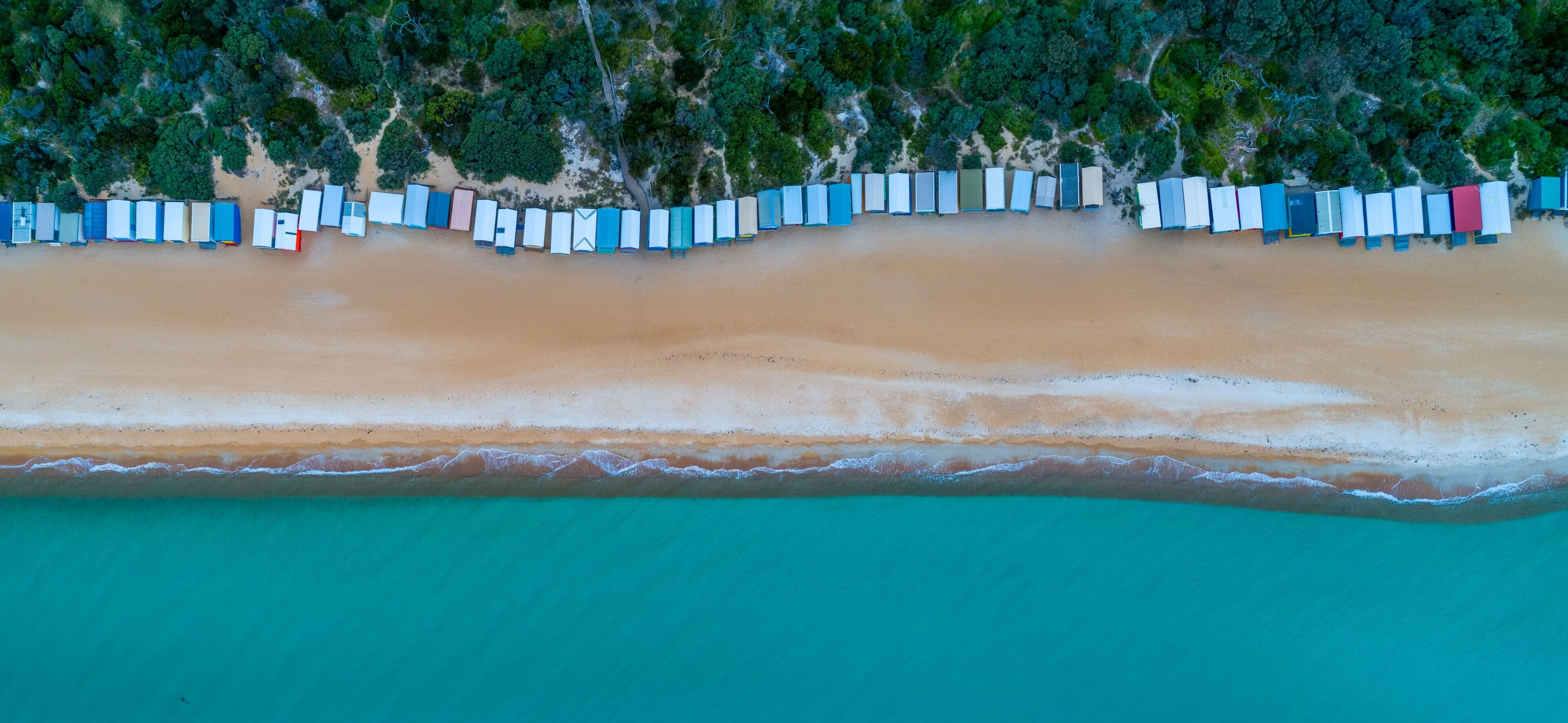 An aerial view of a beach with a row of colorful beach huts along the shoreline and dense forest in the background.