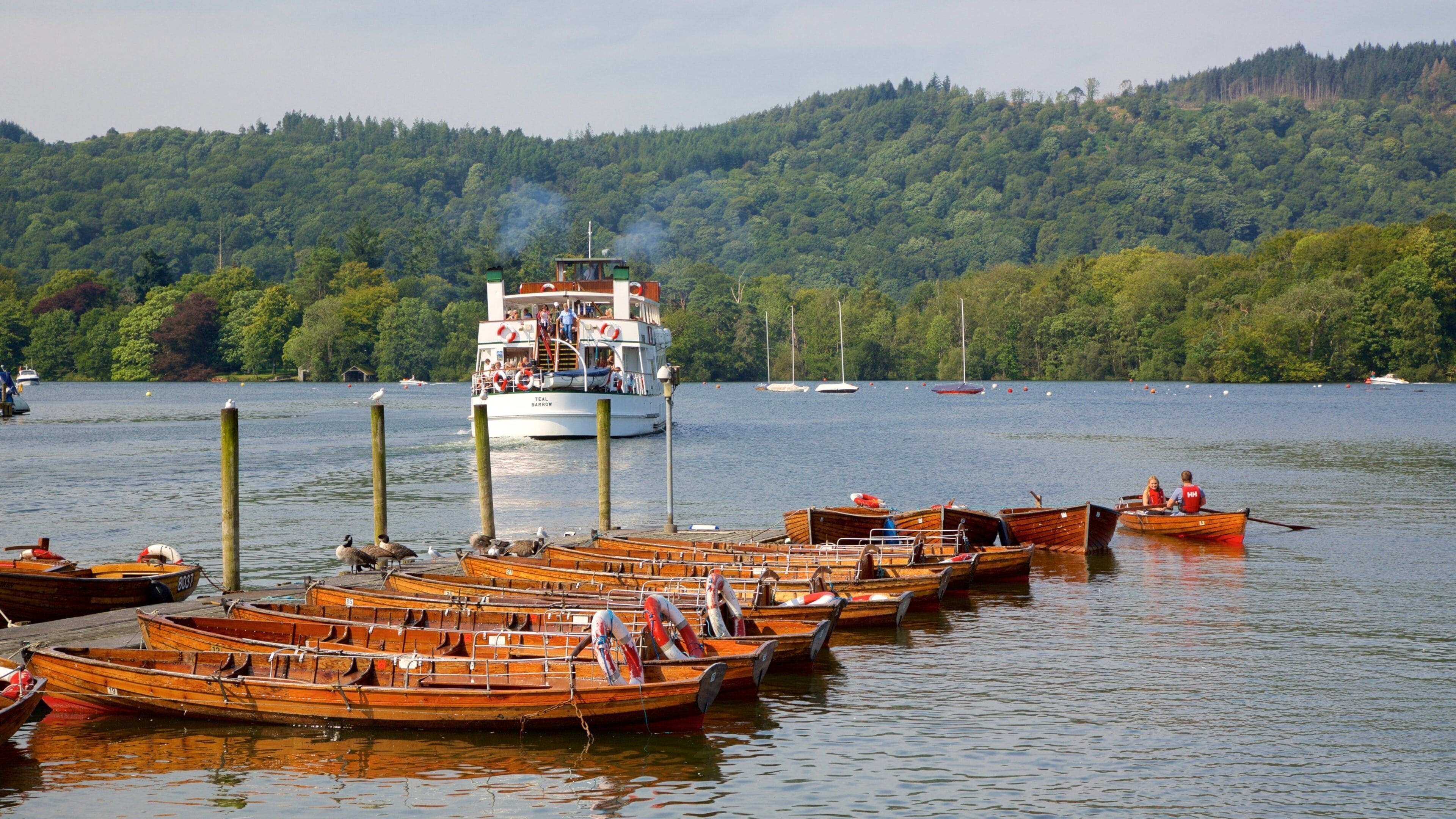 A lake with several wooden boats docked along the shore and a larger boat with people on it in the background.