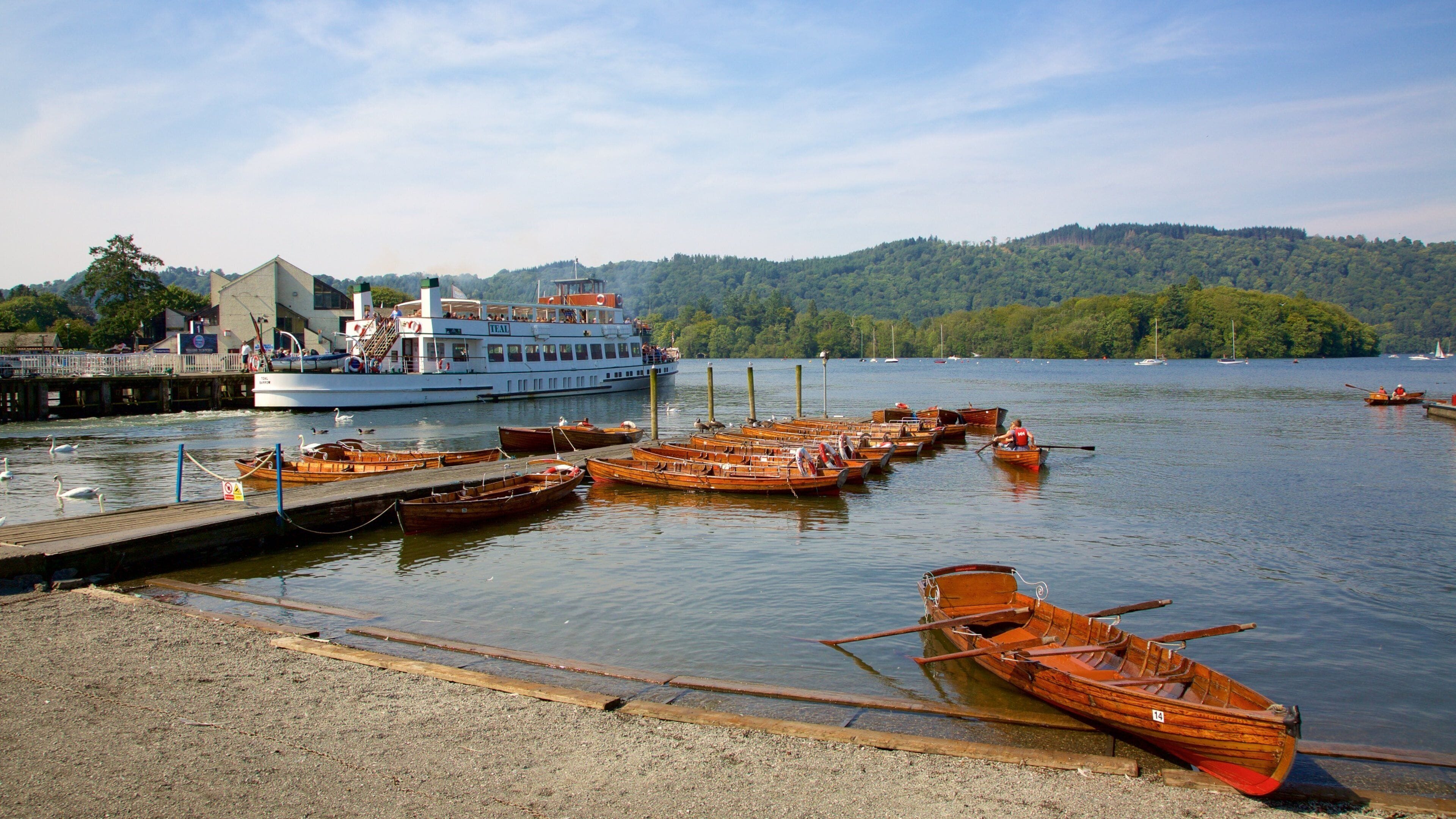 A lake with wooden boats docked at a pier, a large white boat, and a forested hill in the background.