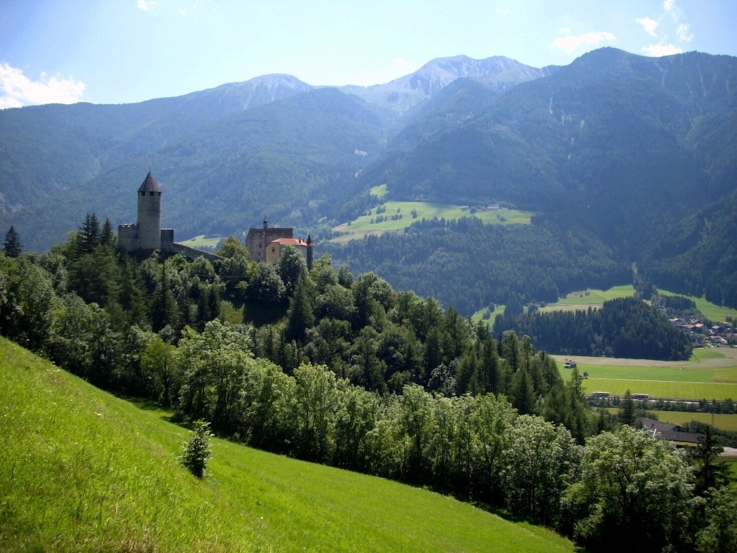 A castle on a hill with a forested valley and mountains in the background.