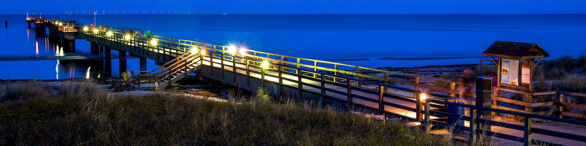 A wooden pier extending into a body of water at dusk, with lights along its length and a small wooden structure at the end.