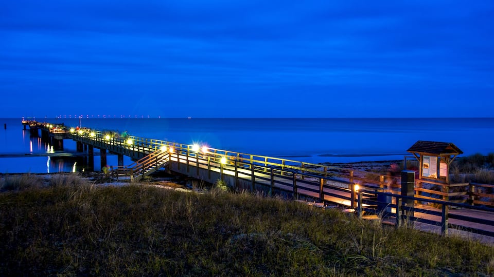 A wooden pier extending into a body of water at dusk, with lights along its length and a small wooden structure at the end.