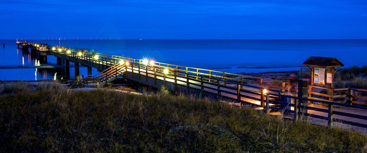A wooden pier extending into a body of water at dusk, with lights along its length and a small wooden structure at the end.