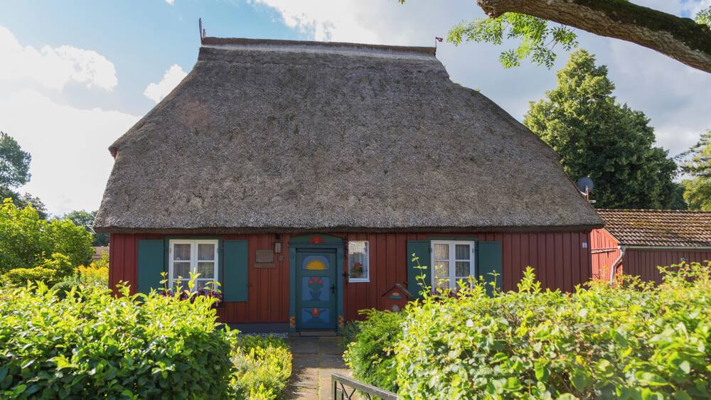 A thatched-roof house with a red exterior and a blue door, surrounded by lush greenery.