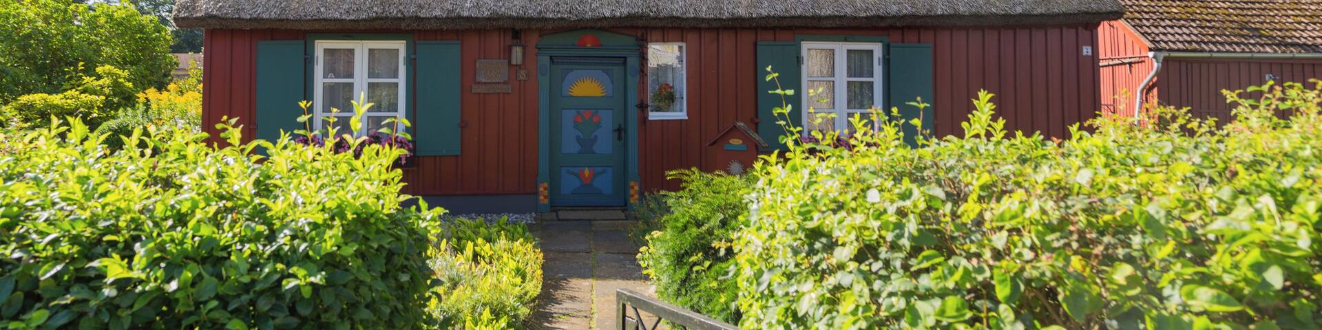 A thatched-roof house with a red exterior and a blue door, surrounded by lush greenery.