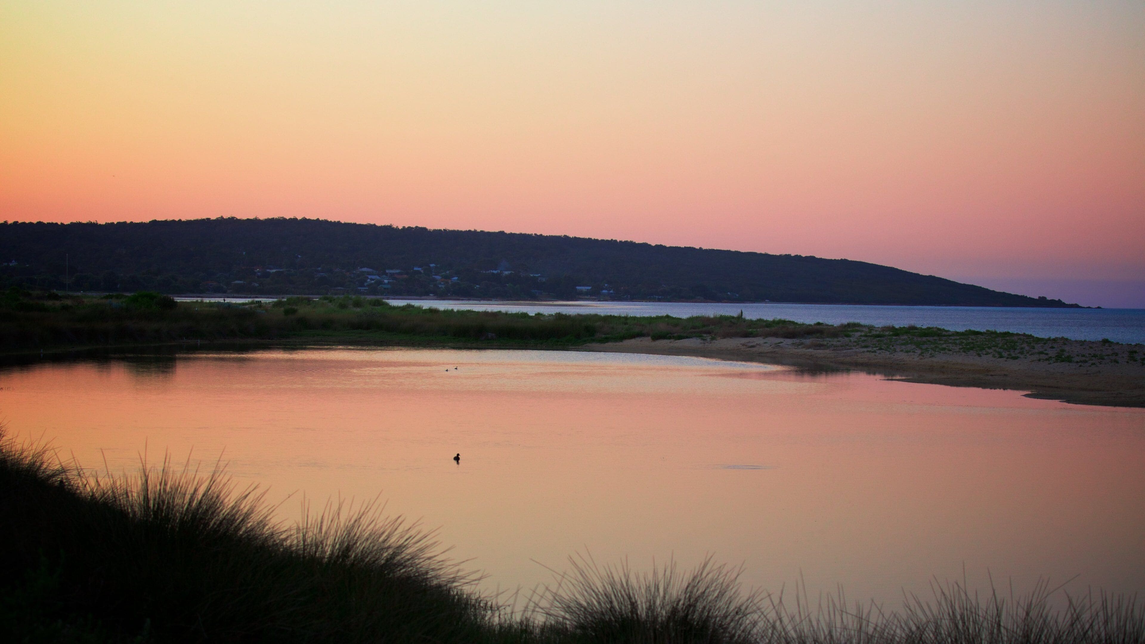 Un lago tranquilo al atardecer, con una colina distante y vegetación escasa.