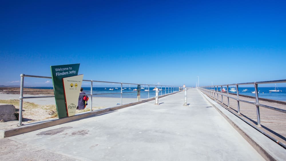 Ein betonierter Wegweiser mit der Aufschrift 'Welcome to Flinders Jetty' führt zu einem Strand mit Booten im Wasser.