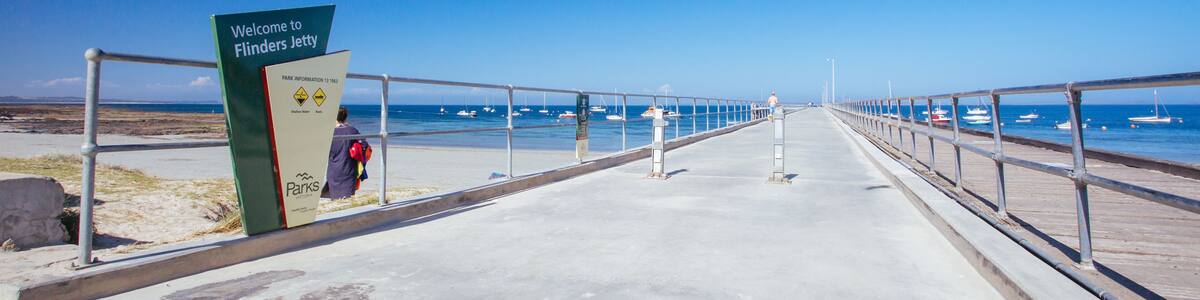 Ein betonierter Wegweiser mit der Aufschrift 'Welcome to Flinders Jetty' führt zu einem Strand mit Booten im Wasser.