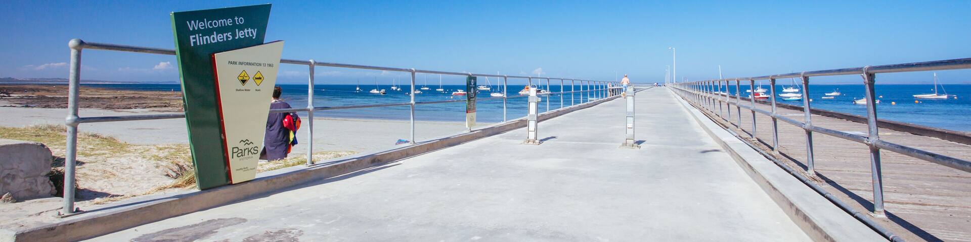 A concrete walkway with a sign reading 'Welcome to Flinders Jetty,' leading to a beach with boats in the water.
