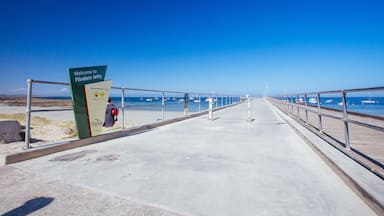 Uma passarela de concreto com uma placa indicando 'Welcome to Flinders Jetty', levando a uma praia com barcos na água.