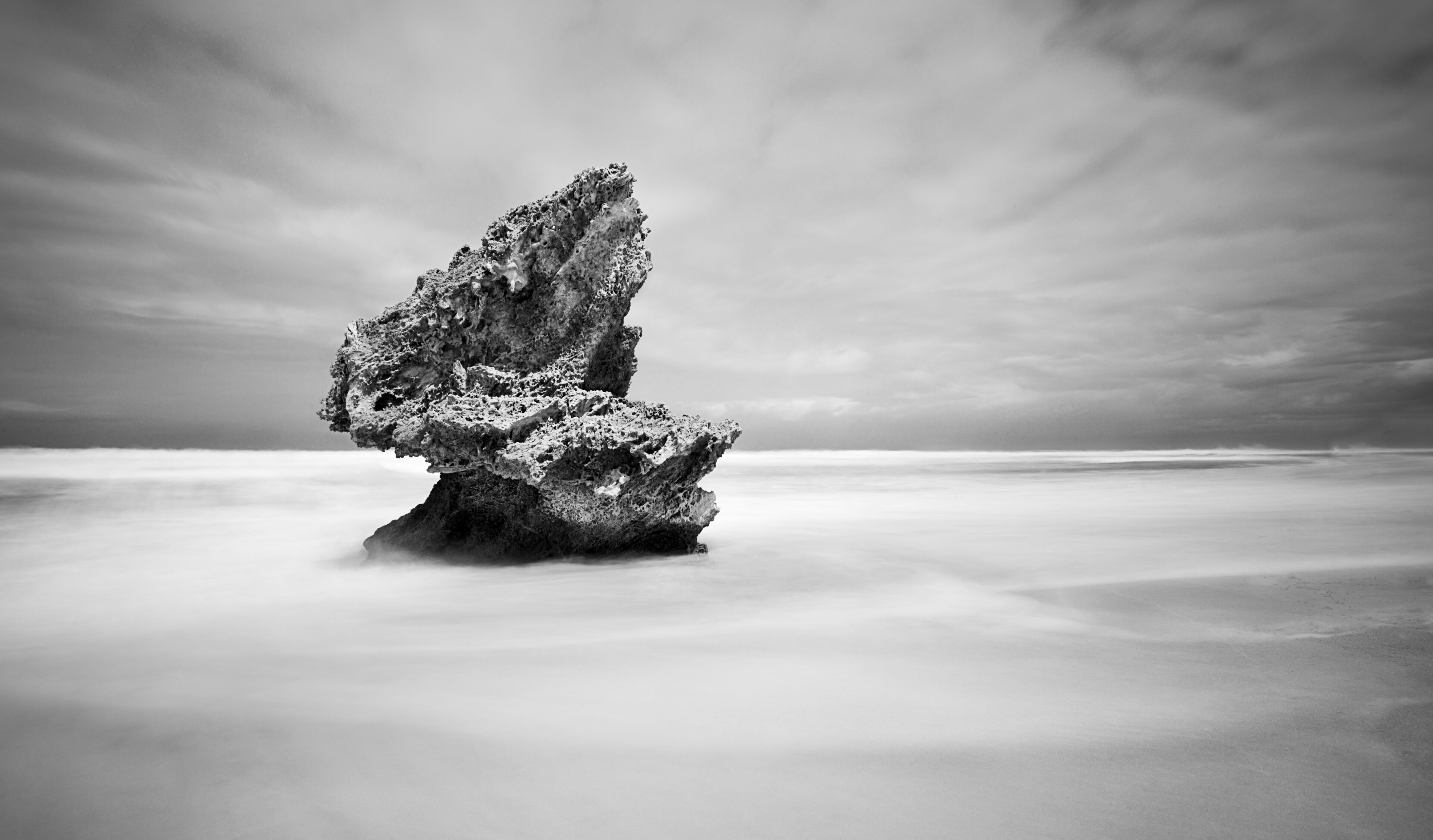 A rock formation in a vast, calm sea under a cloudy sky.