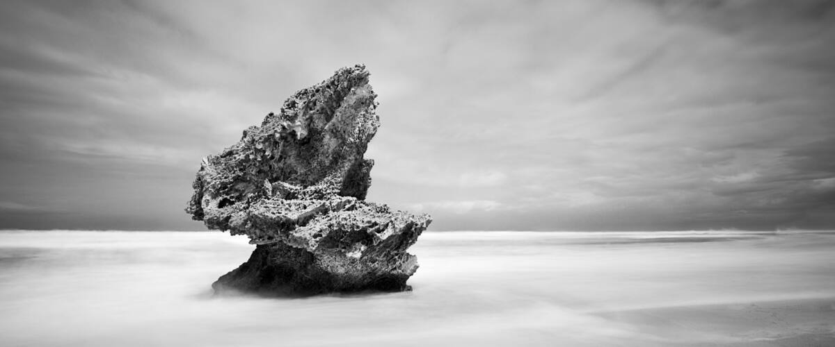 A rock formation in a vast, calm sea under a cloudy sky.