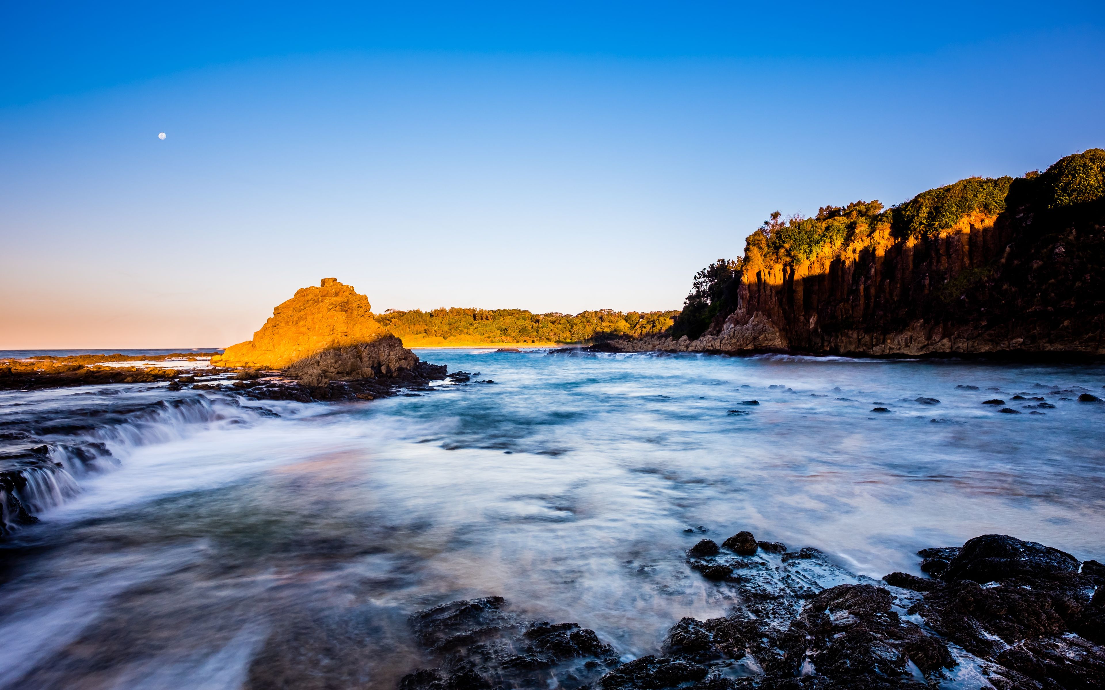 A coastal landscape with a prominent rock formation, a flowing river, and a forested cliff.