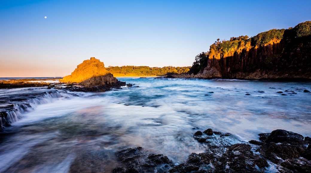 A coastal landscape with a prominent rock formation, a flowing river, and a forested cliff.