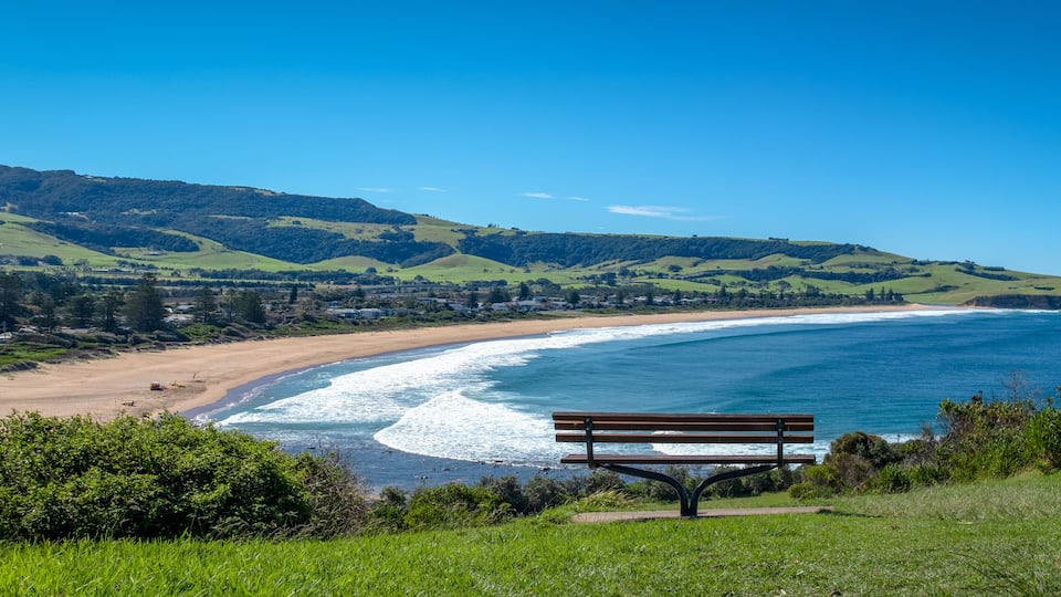 A coastal landscape with a bench overlooking a beach and ocean, surrounded by green hills and clear skies.