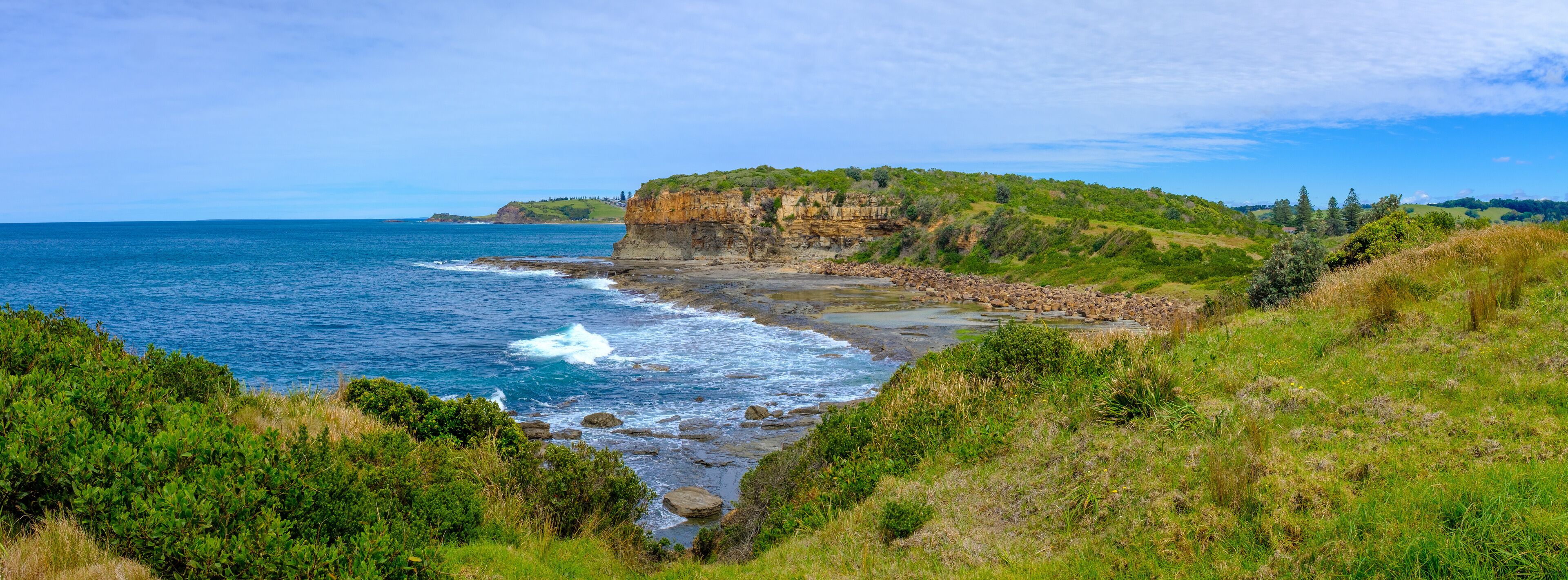 A coastal landscape with a rocky beach, waves crashing onto the shore, and a cliff with vegetation.