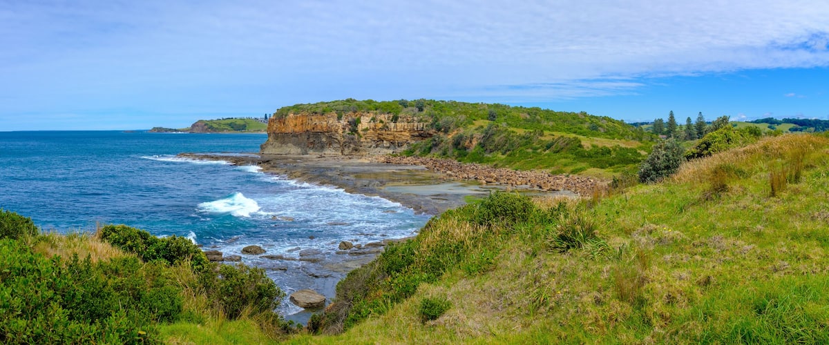 A coastal landscape with a rocky beach, waves crashing onto the shore, and a cliff with vegetation.