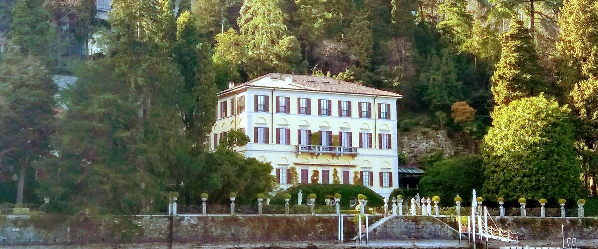 A large white building with red shutters surrounded by trees and a body of water.