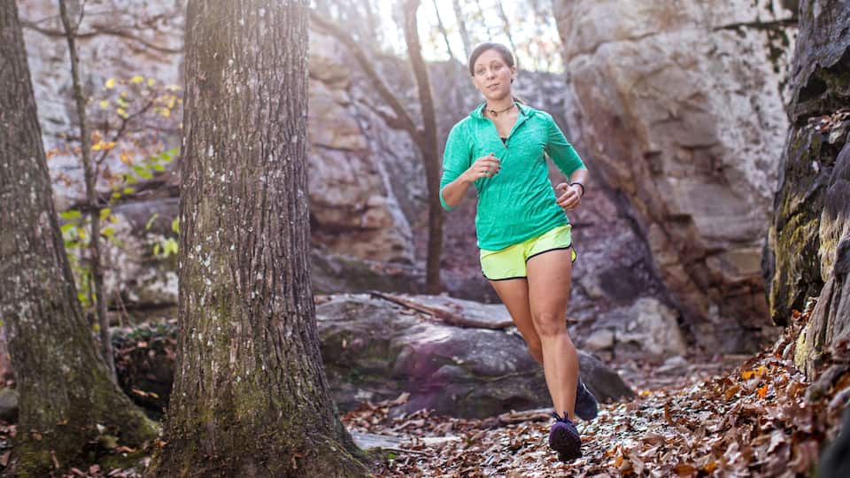 A person running through a forested area with large trees and rocky terrain.