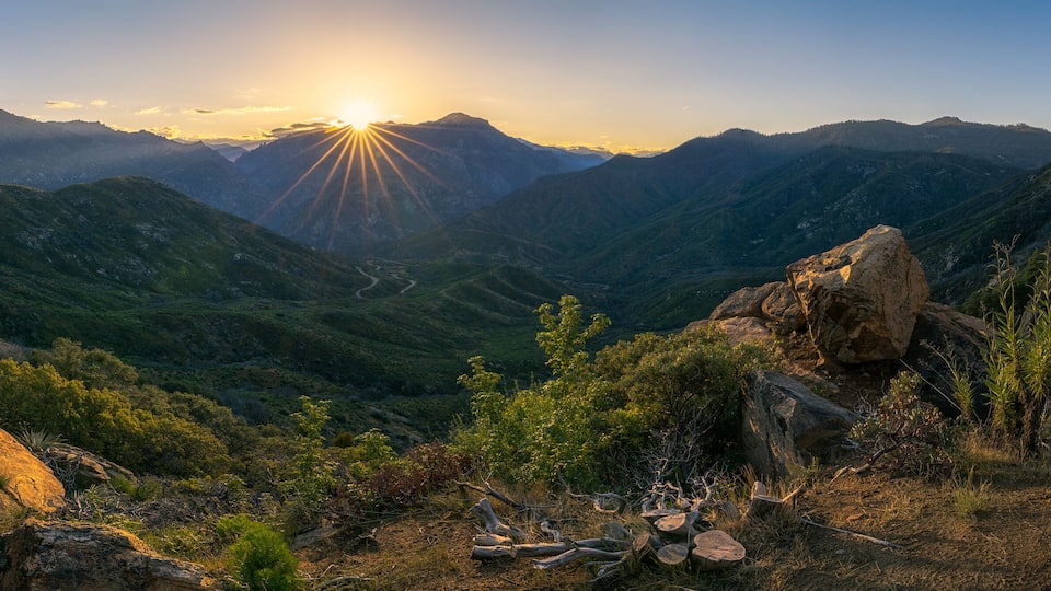 A mountainous landscape with a sunlit valley, rocky outcrops, and dense greenery.