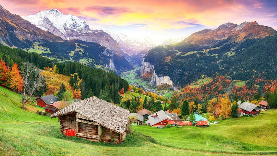 A mountainous landscape with a valley, houses, and autumn-colored trees.