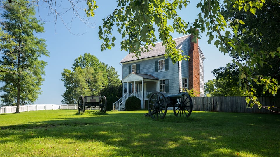 A historic house with a large chimney, surrounded by a wooden fence and two horse-drawn carriages.