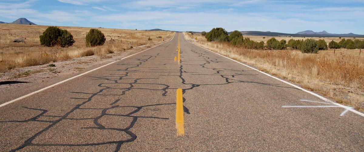 A cracked road in a dry, open landscape with sparse vegetation and distant mountains under a clear sky.