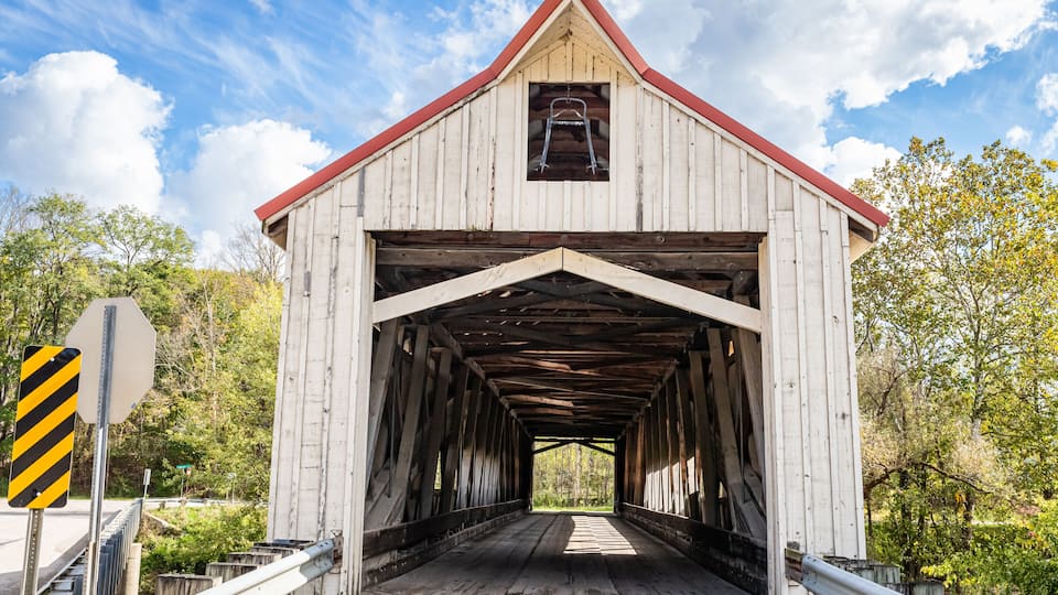 A covered wooden bridge with a red roof, surrounded by trees and a clear sky.