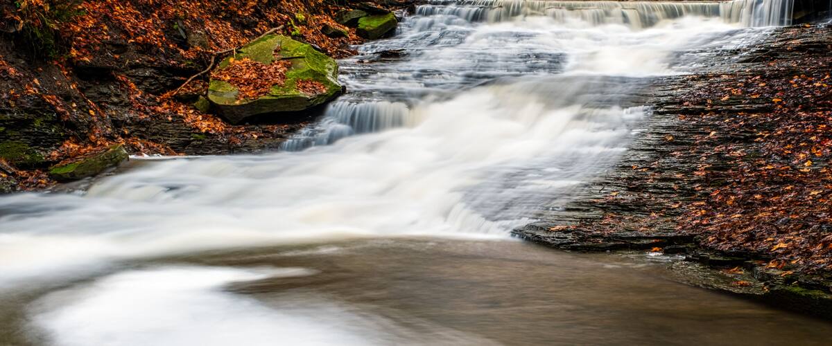 A waterfall cascading over rocks in a forested area with autumn leaves.