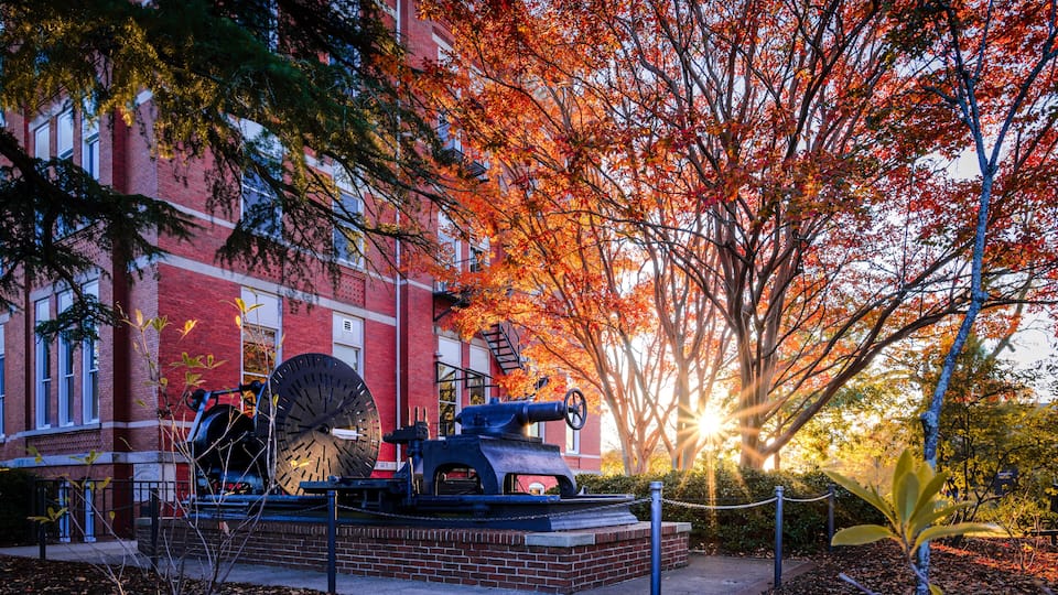 A cannon in front of a red brick building with autumn foliage.