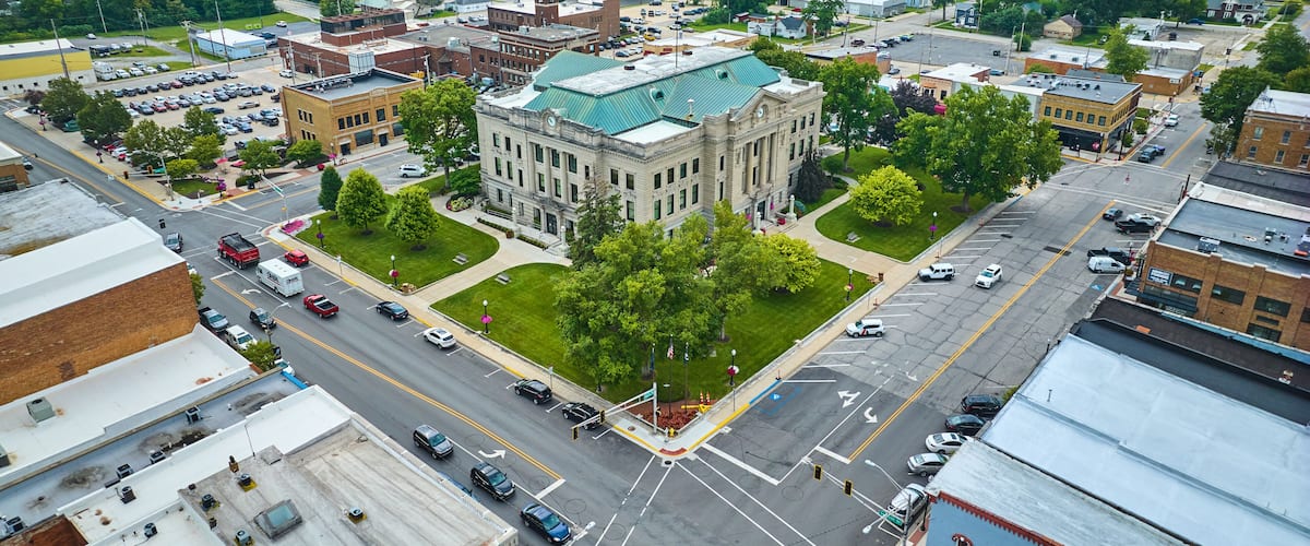 An aerial view of a town with a prominent historic building, a central intersection, and surrounding greenery.