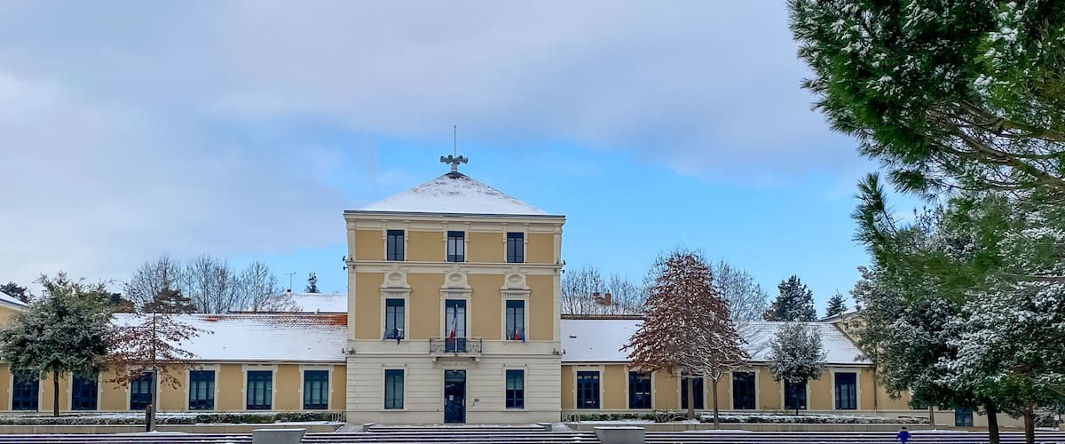 A snow-covered courtyard with a yellow building featuring a central tower and multiple windows.
