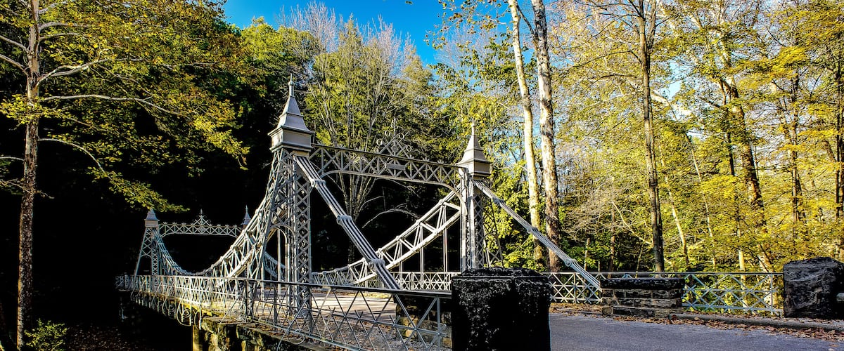 A suspension bridge over a river, surrounded by autumn trees and a stone wall.