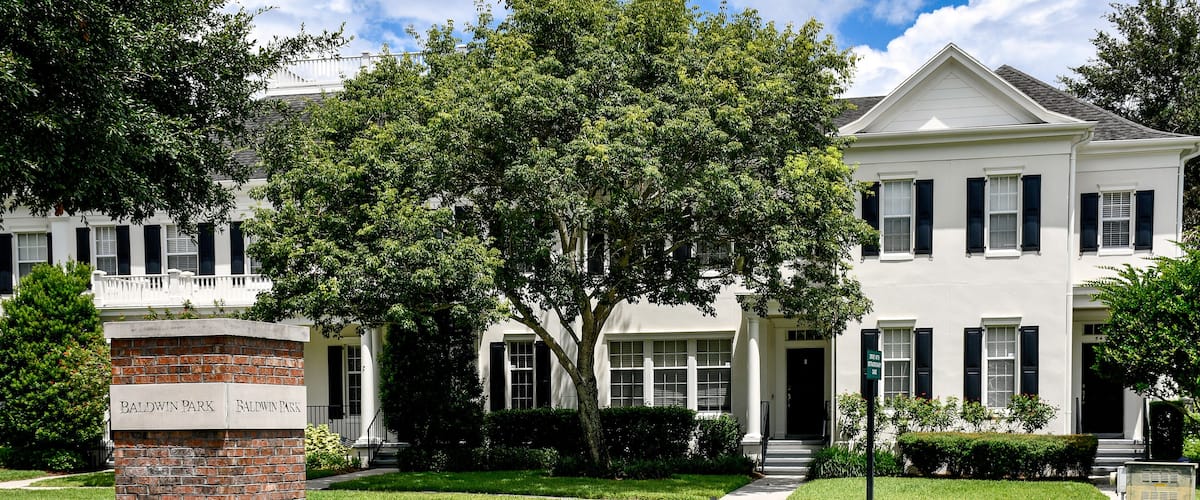 A white house with black shutters, a brick pillar with Baldwin Park sign, and a well-maintained lawn.