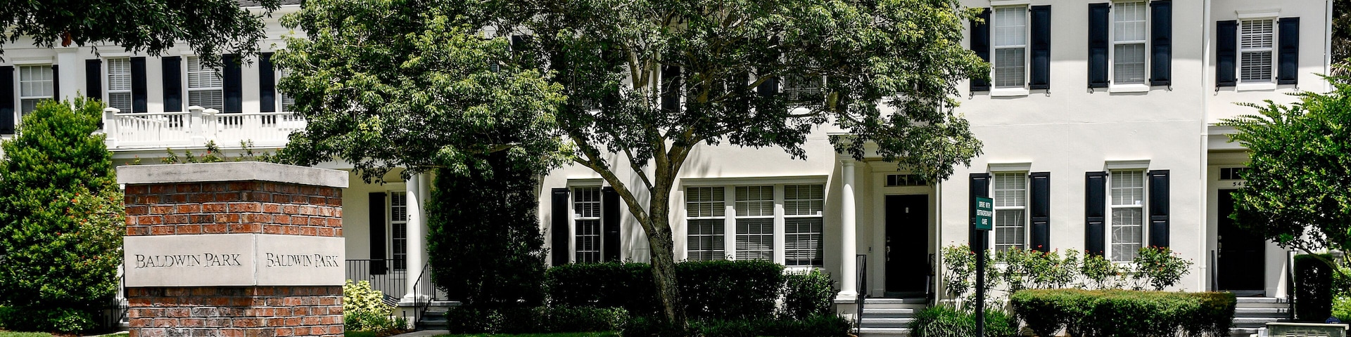 A white house with black shutters, a brick pillar with Baldwin Park sign, and a well-maintained lawn.