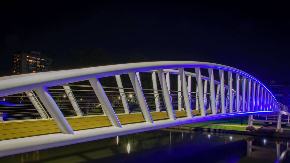 A modern bridge with white railings and blue lighting, spanning over a body of water with a cityscape in the background.