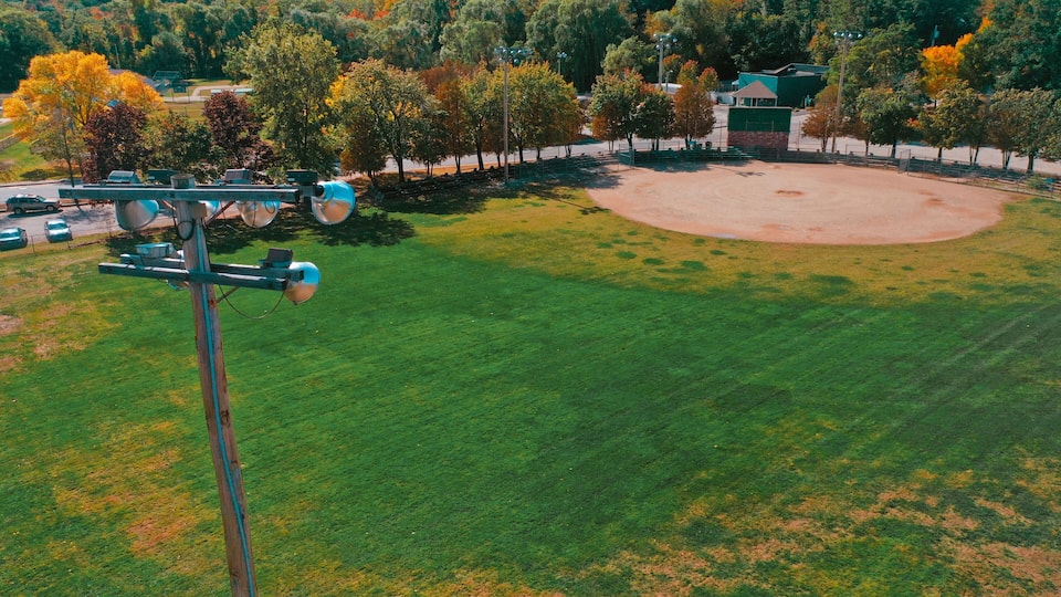 A baseball field with a scoreboard, surrounded by trees and a green building.