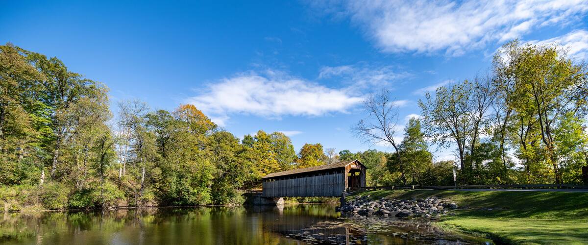 A covered bridge over a calm river with surrounding trees and a clear sky.
